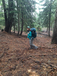 A slightly older version of the same woman, still smiling, wearing a backpack and a turquoise jacket in a forest.