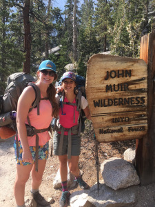 Two women wearing backpacking backpacks, smiling at the signpost to the John Muir Wilderness in California