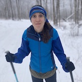 A woman on cross country skis surrounded by snowy trees makes a silly face at the camera.